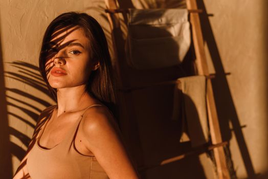 A young woman in a beige bodysuit enjoys the sunbeams in a modern bathroom. Shadows from tropical leaves