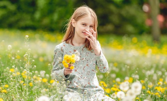 Little girl holding yellow blowballs flowers in hands and sitting in blossom field looking at camera. Cute child kid with dandelions at nature