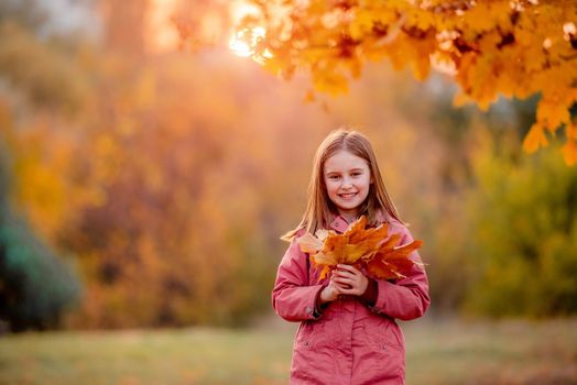 Preteen girl kid holding yellow fallen leaves bouquet in her hands and smilng at autumn park. Beautiful portrait of child outdoors at nature