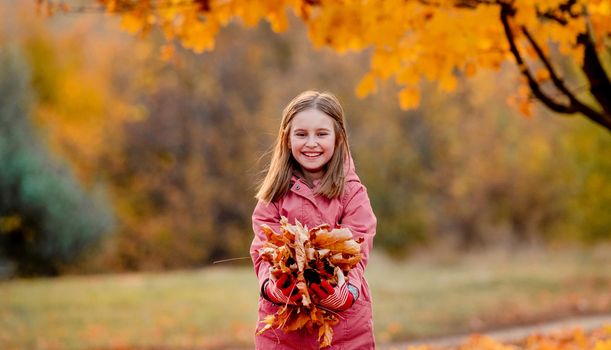 Preteen girl kid holding yellow fallen leaves in hands and smilng at autumn park. Beautiful portrait of child outdoors at nature