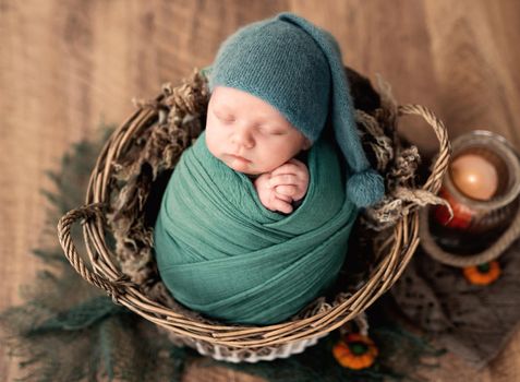 Newborn baby boy swaddled in fabric sleeping in basket decorated with pumpkins. Infant kid napping halloween portrait