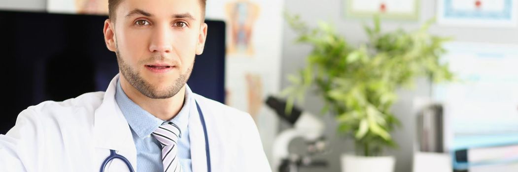 Handsome male doctor sitting at the table in the clinic, close-up, blurry background. Attentive doctor listens, portrait