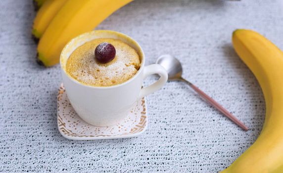 banana cupcake in a mug. Muffin banana cooked in the microwave. Decorated with a cherry. White mug with dessert and fresh banana in the background. High quality photo