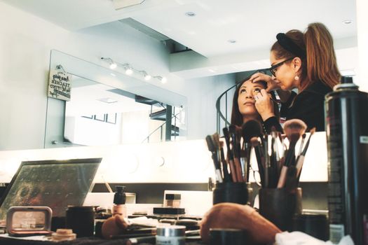 A professional makeup artist in a studio painting a woman's face - people reflected in mirror