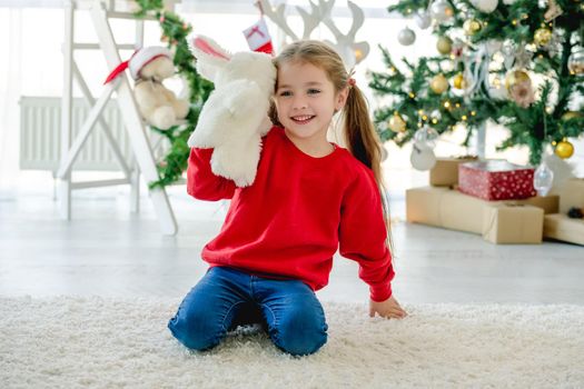 Child girl in Christmas time sitting on carpet in room with decorated tree and gifts. Pretty kid at home in New Year holidays