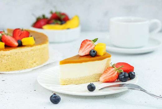 A piece of cheesecake with mango on a plate with a tea fork decorated with berries and flowers on a gray background. Healthy food.