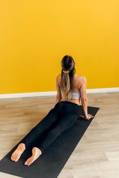 Side view of a beautiful young woman in sportswear practicing yoga on an exercise mat on a yellow background. Stretching the muscles of the back and legs, yoga.