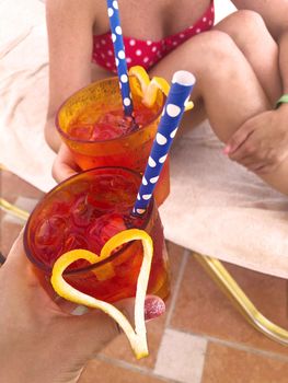 girls holding two cooling, bright, feminine cocktails with hearts. Drinking cocktails by the pool on vacation. Summer holiday concept with alcoholic cocktails in hands. clinking glasses by the pool