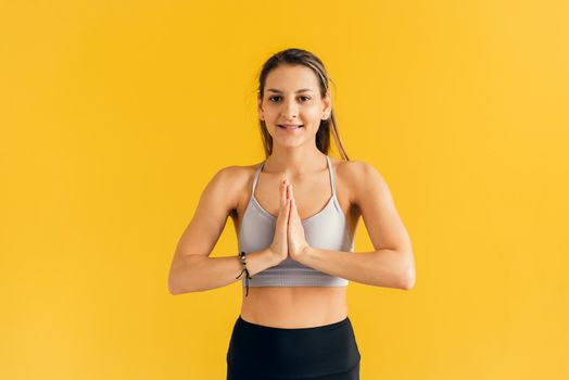 Concentrated young woman in tight sportswear practicing yoga, holding hands in namaste gesture and meditating, relaxing, looking at camera. Woman praying and holding hands in zen on yellow background.