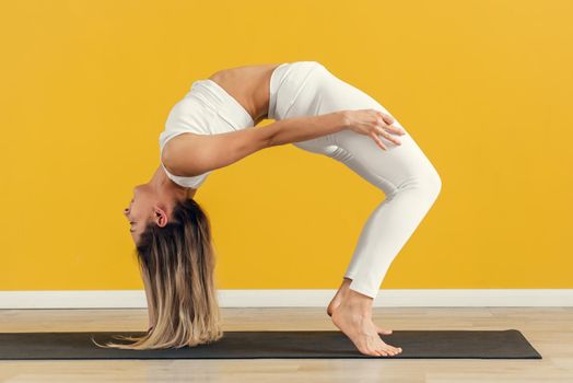 Young attractive yogi woman practicing bridge pose without arms, stretching in Urdhva Dhanurasana exercise on yellow background.