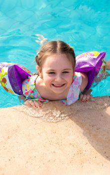 baby girl floating in the pool inflatable over-sleeves. holding on to the curb of the swimming pool, with blue transparent water. the child in the pool smiles widely. summer background