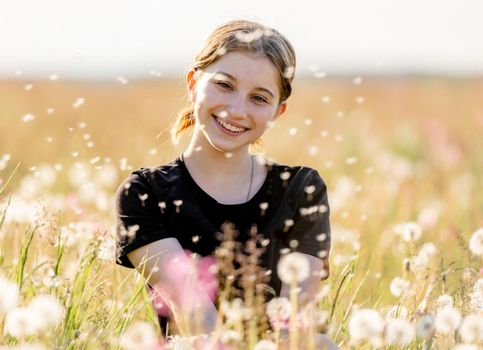 Pretty girl teenager sitting in field with flying dandelions and smiling. Beautiful young female person model at nature in sunny day portrait