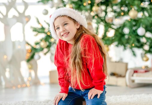 Little girl child wearing Santa hat in Christmas time sitting and smiling at decorated New Year home with festive tree. Cute kid in Xmas time