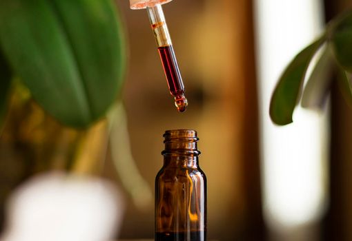 a drop of serum with a pipette drips over a brown glass jar on green background close-up. selective focus. beauty concept. natural cosmetic. Daily skin care. Anti-aging serum with natural ingredients