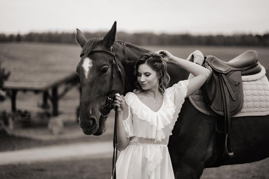 A girl in a white sundress stands next to a brown horse in a field in summer.black and white photo.