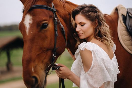 A girl in a white sundress stands next to a brown horse in a field in summer.