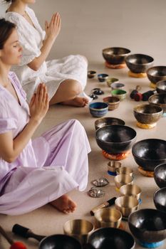 Two women are sitting with Tibetan bowls in the lotus position before a yoga class in the gym.