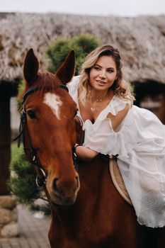 A woman in a white sundress riding a horse near a farm.