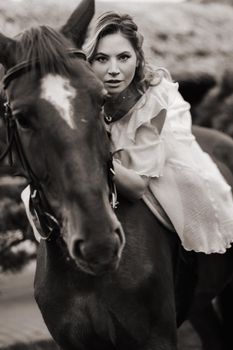 A woman in a white sundress riding a horse near a farm. black and white photo.