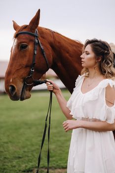 Beautiful girl in a white sundress next to a horse on an old ranch.