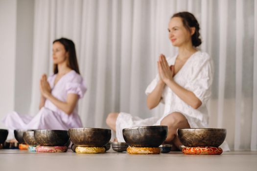 Two women are sitting with Tibetan bowls in the lotus position before a yoga class in the gym.