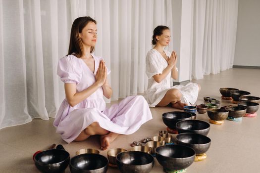 Two women are sitting with Tibetan bowls in the lotus position before a yoga class in the gym.