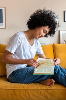 Young African American woman reading a book sitting on yellow couch. Female relaxing at home. Vertical image. Lifestyle concept.