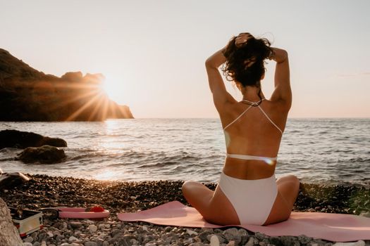 Young woman in swimsuit with long hair practicing stretching outdoors on yoga mat by the sea on a sunny day. Women's yoga fitness pilates routine. Healthy lifestyle, harmony and meditation concept.