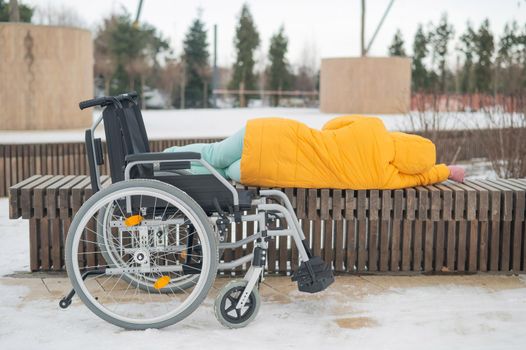 Homeless woman sleeping on a park bench next to a wheelchair