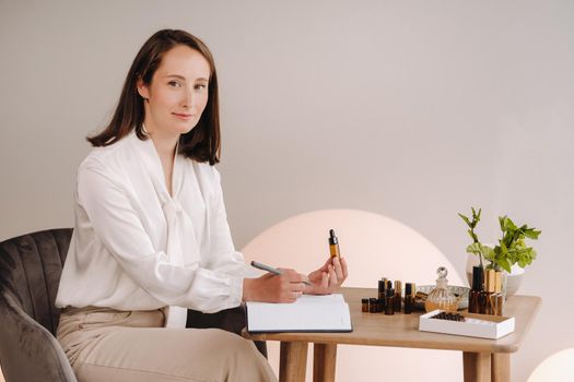 The aromatherapist girl is sitting in her office and holding a bottle of aromatic oil in her hands and writing something down. there are essential oils on the table.