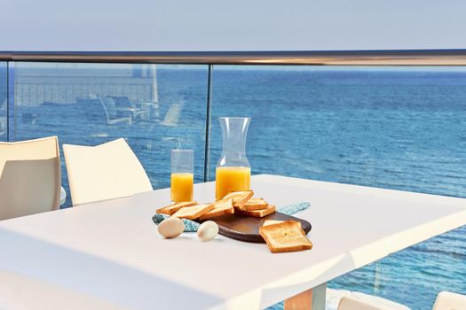 Table for two on outdoor hotel balcony with a sea view. European vacation breakfast, food selfie. Concept of a rest and healthy nutrition. There are toasts on a wooden tray, eggs, orange juice.