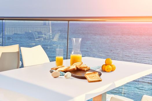 Table for two on outdoor hotel balcony with a sea view. European vacation breakfast, food selfie with a sunflare. Concept of a rest and healthy nutrition. There are toasts, eggs, orange juice.