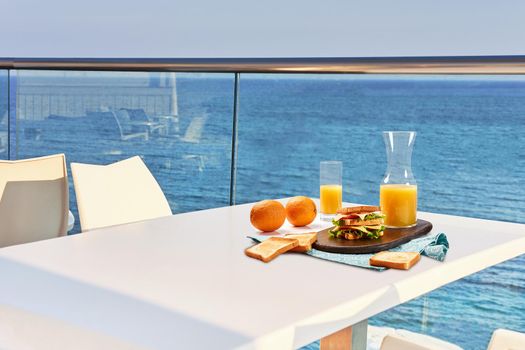 Table for two on outdoor hotel balcony with a sea view. European vacation breakfast, food selfie. Concept of a rest and healthy nutrition. There are toasts, sandwich on a wooden tray, orange juice.