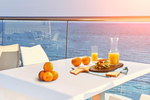 Table for two on outdoor hotel balcony with a sea view. European vacation breakfast, food selfie with a sunflare. Concept of a rest and healthy nutrition. There are toasts, sandwich, orange juice.