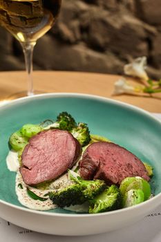 Close-up photo of a tasty roasted meat with broccoli and zucchini seasoned with sauce. Served on a blue deep plate, with a glass of wine. Restaurant cooking concept. Side view.