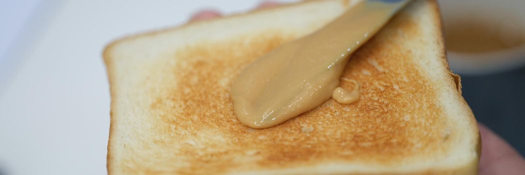 In hands toasted toast with peanut butter, close-up. Spread the nut butter with a knife, breakfast. Snack at home