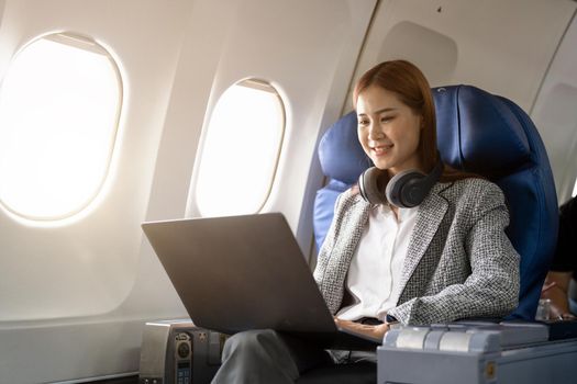 Asian young woman wearing headphone using laptop sitting near windows at first class on airplane during flight, Traveling and Business concept.