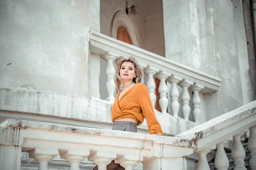 A middle-aged woman looks like a good blonde with curly beautiful hair and makeup on the background of the building. She is wearing a yellow sweater