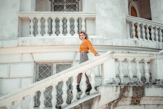 A middle-aged woman looks like a good blonde with curly beautiful hair and makeup on the background of the building. She is wearing a yellow sweater