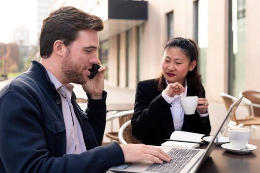 caucasian businessman talking by phone while has a work meeting over coffee with his asiatic partner on a terrace in the financial district, concept of entrepreneurship and business