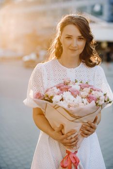 A happy woman in a white dress at sunset with a bouquet of flowers in the city.