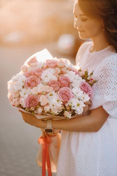 A happy woman in a white dress at sunset with a bouquet of flowers in the city.