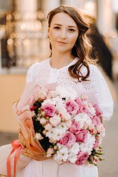 A happy woman in a white dress at sunset with a bouquet of flowers in the city.