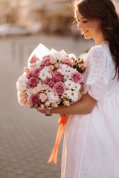 A happy woman in a white dress at sunset with a bouquet of flowers in the city.