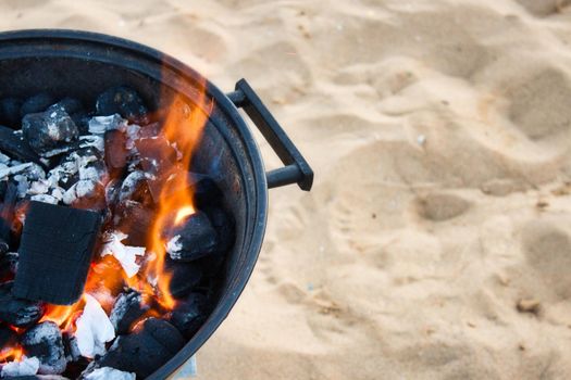 Closeup on charcoal burning with a yellow flame in a round metallic barbeque on a sandy beach