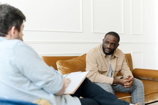 An irritated African American emotionally talks about problems to a psychologist at a session, a confused unhappy young man undergoes therapy with a psychiatrist