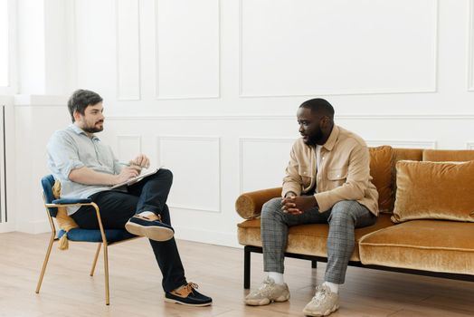 The therapist takes notes while talking to a depressed male patient and talks to an African American patient. Desperate black man suffering from nervous breakdown or depression having a session with a psychologist.