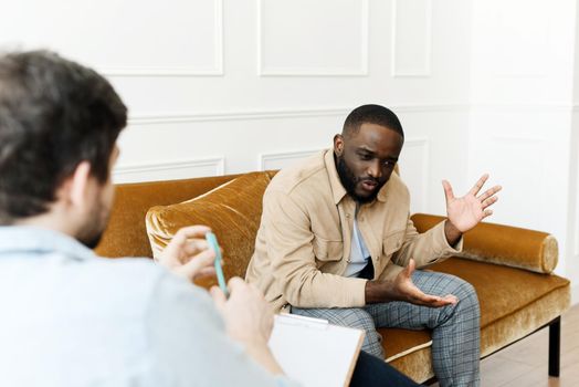 An irritated African American emotionally talks about problems to a psychologist at a session, a confused unhappy young man undergoes therapy with a psychiatrist