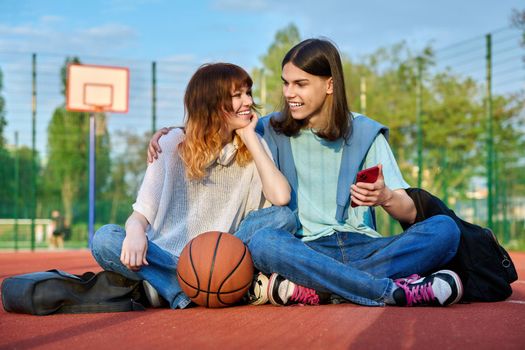 Couple of teenage student friends sitting on outdoor basketball court, with ball backpacks, laughing, talking, looking in smartphone. Youth, active healthy lifestyle, education, urban sport concept