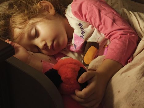 Adorable little girl asleep in her bed dimly lit by a bedside table lamp with a warm bulb